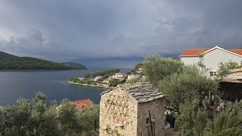 Dunkle Regenfront über der Bucht, im Vordergrund Olivenbäume und Steinhäuser